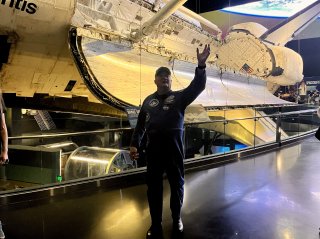 Former astronaut Bruce Melnick wearing a blue flight suit and standing in front of the space shuttle Atlantis on display at the space center.
