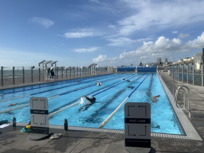 An outdoor 50-meter pool with six lanes which are quite busy with swimmers. The sky is blue, but clouds are building on the horizon.